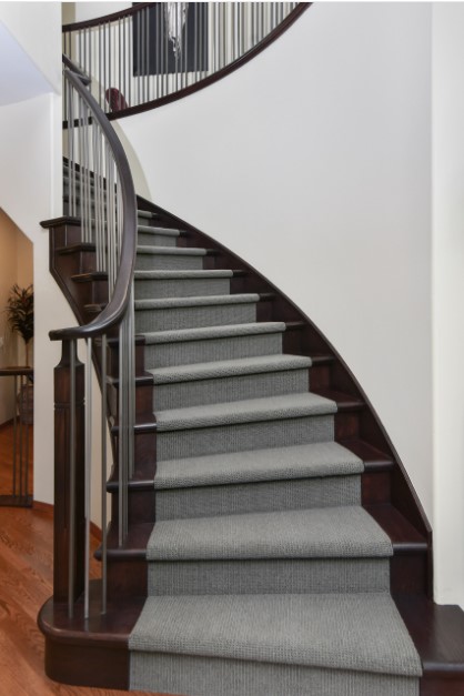 Dark wood curved staircase with thin guard rails and white walls. There is a grey carpeted stair runner along the middle. The design is carried through to the upper level.